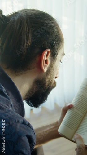 Vertical video. A bearded man is sitting on the couch near a window, engrossed in reading a book. Calm and tranquility, with natural light streaming through sheer curtains.