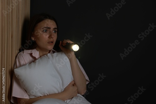 Fear of darkness. Scared young woman with flashlight hugging pillow indoors at night, space for text photo