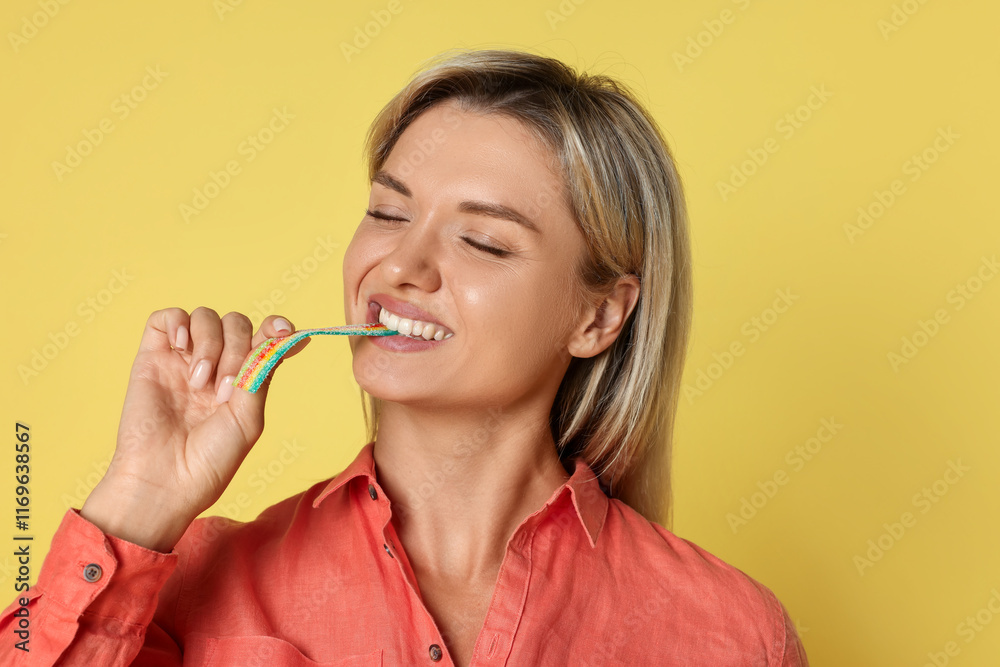Young woman eating tasty rainbow sour belt on yellow background Young woman eating tasty rainbow sour belt on yellow background