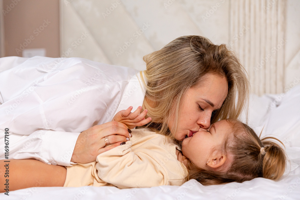 Happy young mother with baby are lying at home in white bedroom. Mom hugs and kisses her little daughter in bed. Mother and daughter are resting in bed at home. Good morning Happy young mother with baby are lying at home in white bedroom. Mom hugs and kisses her little daughter in bed. Mother and daughter are resting in bed at home. Good morning