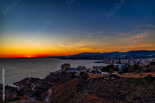 A stunning sunset casts a warm glow over a coastal city, with the sun reflecting beautifully on the calm sea