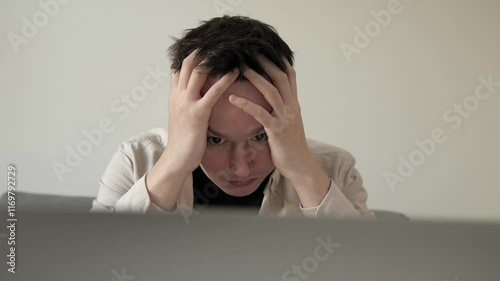 Young man feeling stressed while working on laptop in a modern home interior. Disappointed man covering face palm thinking. Depressed businessman looking frustrated.