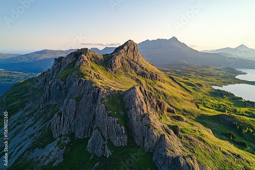 The Old Man of Storli, the most famous peak on Skye island in Scotland, is seen from above during golden hour. The rugged mountain stands tall against a blue sky and lush green landscape with lake