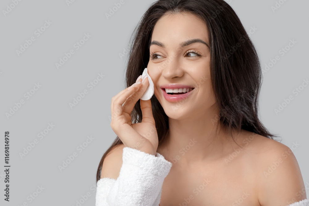Fototapeta premium Beautiful young happy woman in bathrobe with cotton pad on grey background