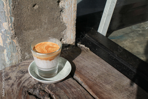 Dirty coffee latte in glass under sunlight and shadow on wooden board. Espresso layer on the top and chilled cold milk or cream at the bottom.
