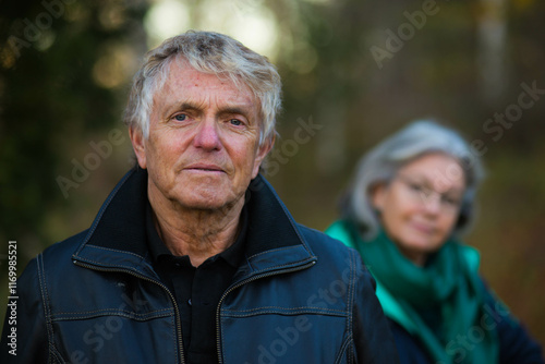 A portrait of a confident old man and his wife, out of focus, in the background