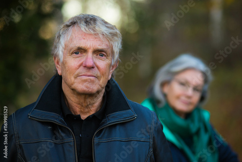 A portrait of an older Swedish man looking into the camera and his wife out of focus in the background