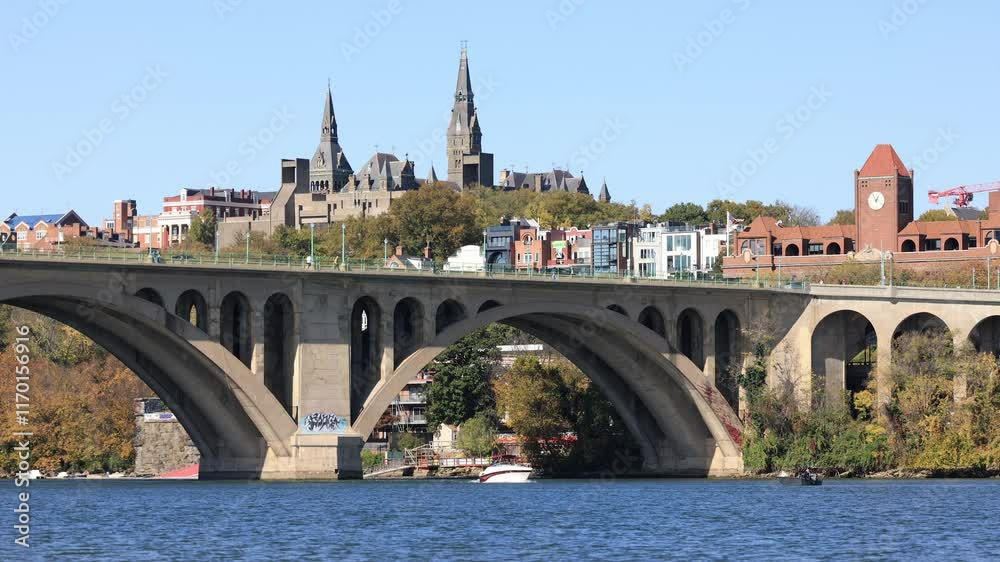 Key Bridge spans the Potomac River, linking D.C. and Rosslyn, Virginia. A motorboat glides below as vehicles, pedestrians, and cyclists cross the busy span on a sunny, fall day.
