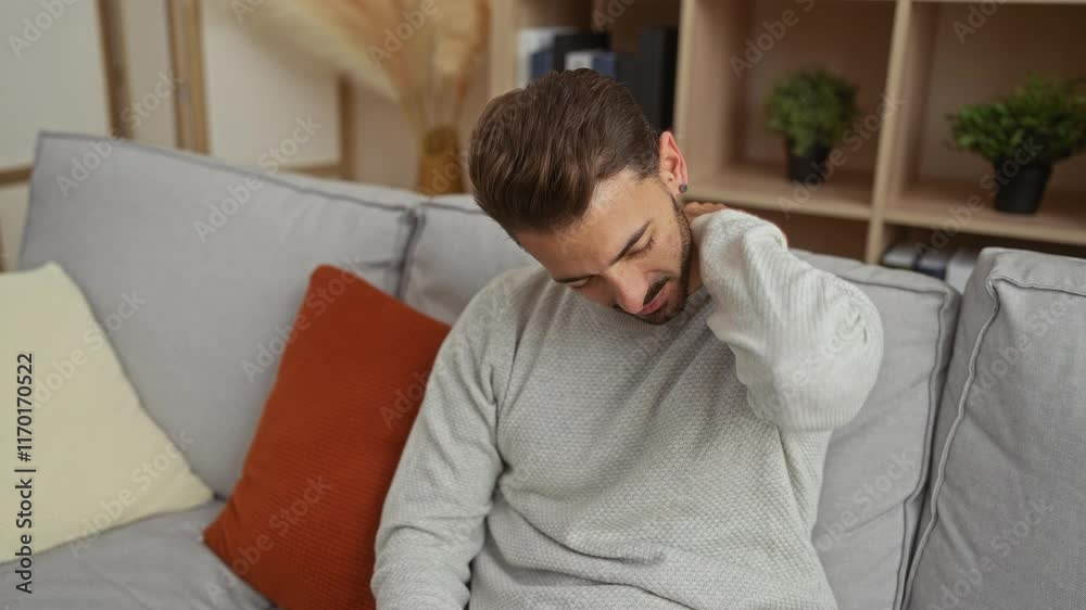 Young man with neck pain sitting on a cozy living room sofa in a home setting, surrounded by cushions and plants, conveying discomfort and a need for relief. Young man with neck pain sitting on a cozy living room sofa in a home setting, surrounded by cushions and plants, conveying discomfort and a need for relief.