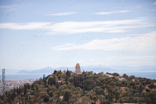 Scenic view of the ancient Tower of the Winds on the Hill of the Nymphs in Athens, Greece. The city sprawls below, with the Aegean Sea and distant mountains visible under a partly cloudy sky photo