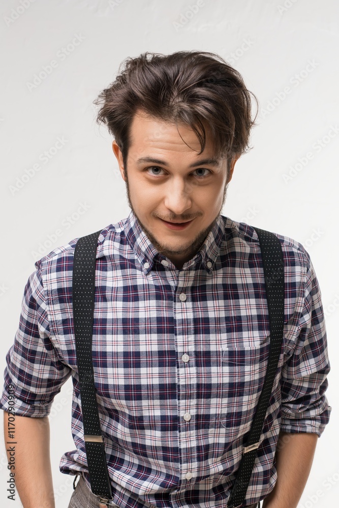 Young man dressed in plaid shirt and suspenders poses against plain background Young man dressed in plaid shirt and suspenders poses against plain background