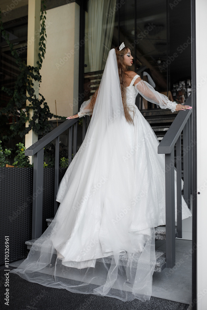 White dress with long transparent sleeves. A woman holds on to the railing of the stairs that lead up. Plants and glass doors or windows are visible in the background. White dress with long transparent sleeves. A woman holds on to the railing of the stairs that lead up. Plants and glass doors or windows are visible in the background.