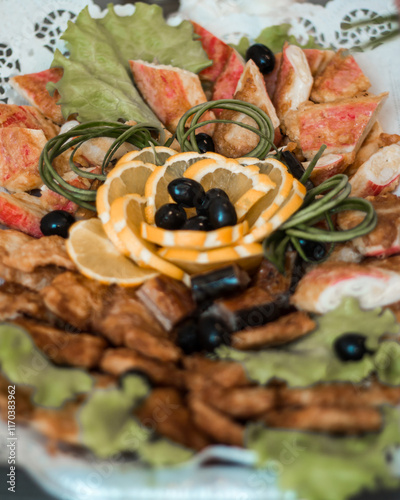 Intricately arranged platter featuring fried items, crab sticks, lettuce, black olives, and a carved lemon flower on a lace like tablecloth. photo