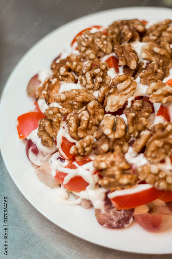 A white plate on a metallic surface holds halved walnuts, sliced red tomatoes, and grapes, topped with a drizzle of creamy white dressing.