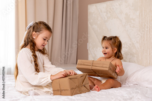 Two children, little sisters sitting on a bed with white bedding, opening gift boxes wrapped in kraft paper. Holidays concept. High quality photo. photo