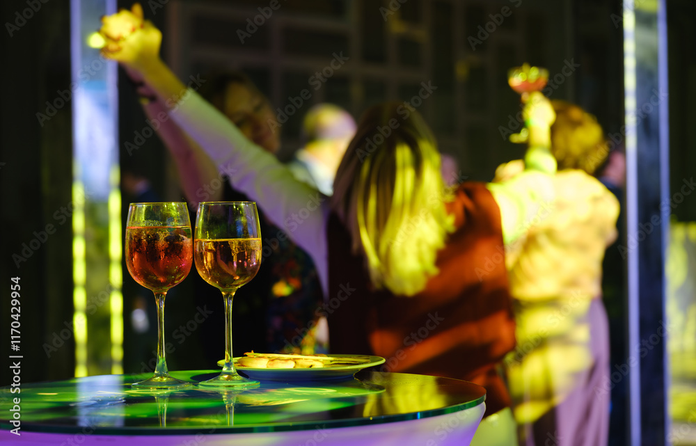 Obraz premium Colorful party scene with wine glasses in foreground, friends dancing and celebrating in vibrant lighting.