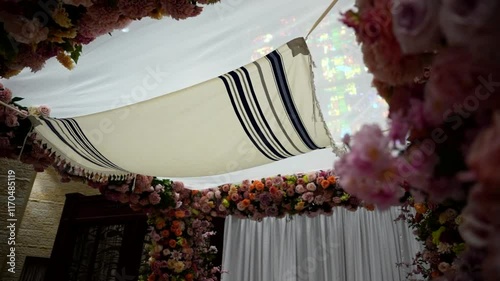 Beautiful indoor chuppah adorned with vibrant pink and orange roses, white fabric canopy, and floral details. Set against a stone wall, perfect for Jewish wedding themes and event inspiration photo