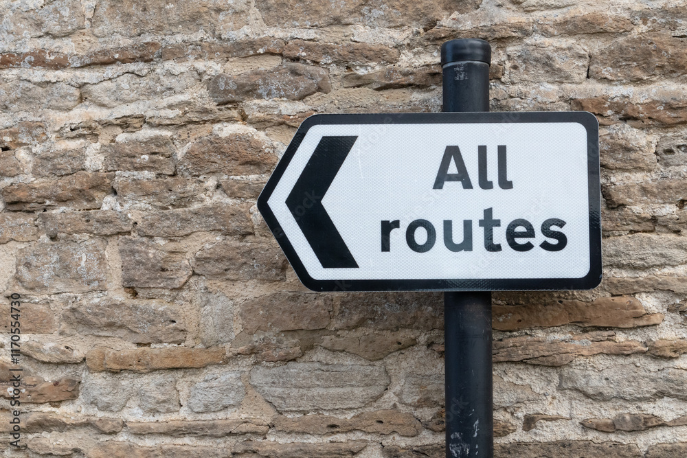 All Routes Road Sign pointing left in front of an old stone wall All Routes Road Sign pointing left in front of an old stone wall