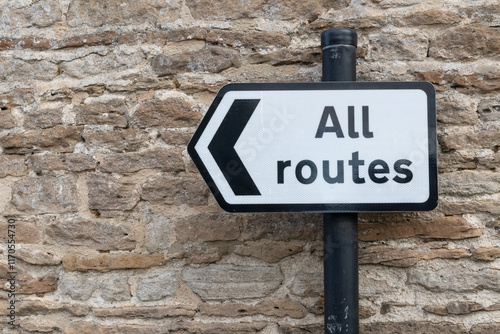 All Routes Road Sign pointing left in front of an old stone wall