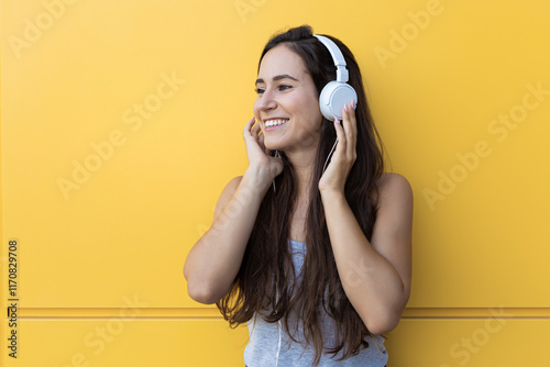Isolated portrait of a beautiful young woman smiling and listening to the music with white headphones in front of a yellow wall
