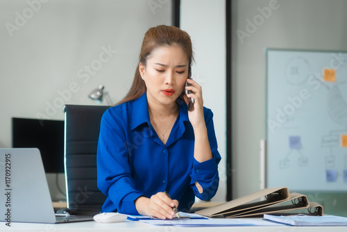 An Asian businesswoman in a blue formal shirt is working at her desk, analyzing financial charts on a laptop and mobile phone, focusing on credit management and financial strategies. photo