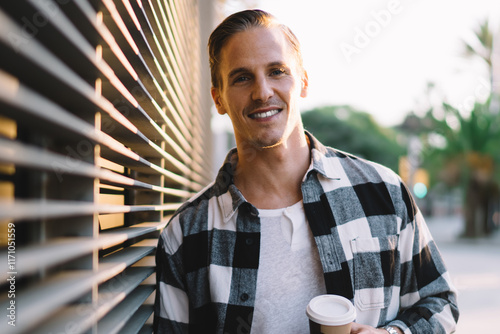 Half length portrait of cheerful caucasian male dressed in casual trendy outfit holding coffee cup enjoying leisure in city,handsome smiling hipster guy looking at camera standing on urban settings photo