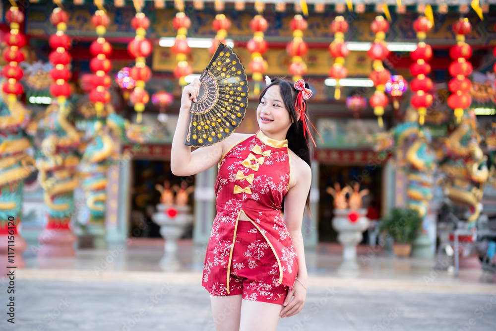 Portrait beautiful cute smiles Asian young woman wearing red traditional Chinese cheongsam decoration and holding a Chinese Fanning for Chinese New Year Festival at Chinese shrine in Thailand