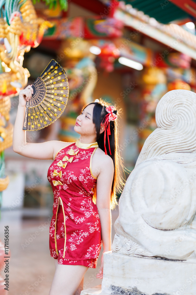 Portrait beautiful cute smiles Asian young woman wearing red traditional Chinese cheongsam decoration and holding a Chinese Fanning for Chinese New Year Festival at Chinese shrine in Thailand
