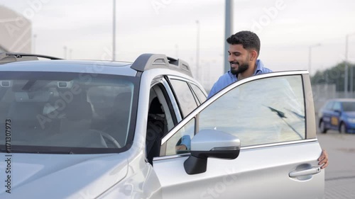 Smiling male commuter wearing shirt, getting in car ready to go to work in morning. Side view of happy bearded man sitting into driver's seat in glossy car parked in city. Concept of driving, commute.