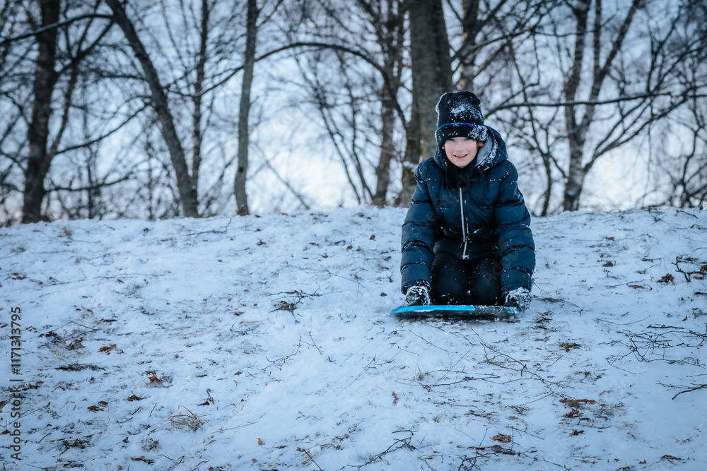 A smiling boy in a black winter jacket and hat sleds downhill on a snow-covered slope. Bare trees in the background add to the wintry atmosphere. A smiling boy in a black winter jacket and hat sleds downhill on a snow-covered slope. Bare trees in the background add to the wintry atmosphere.