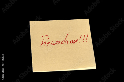 Yellow adhesive paper, similar to a Post-it note, on a black background with the words “Recuerdame in Spanish and three exclamation marks handwritten in red ink.
