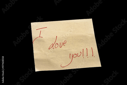 Yellow adhesive paper, similar to a Post-it note, on a black background with the word “I love You” handwritten in red ink.