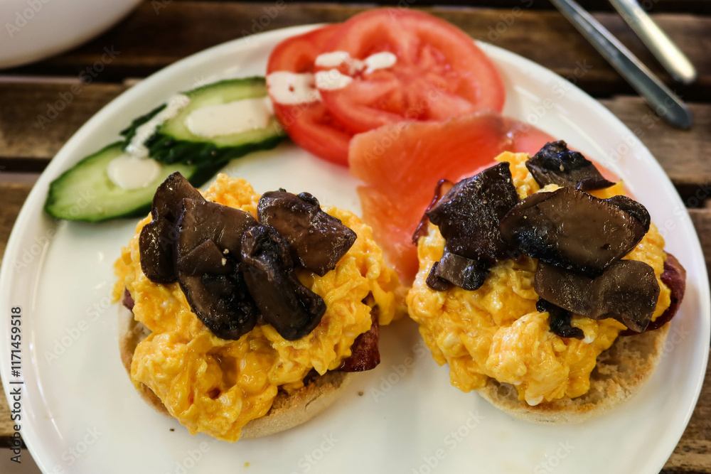 close up of American breakfast with scramble egg on muffin bread in half and stir fried mushroom. uncooked tomato and cucumber on the side on a white plate on a wooden table close up of American breakfast with scramble egg on muffin bread in half and stir fried mushroom. uncooked tomato and cucumber on the side on a white plate on a wooden table