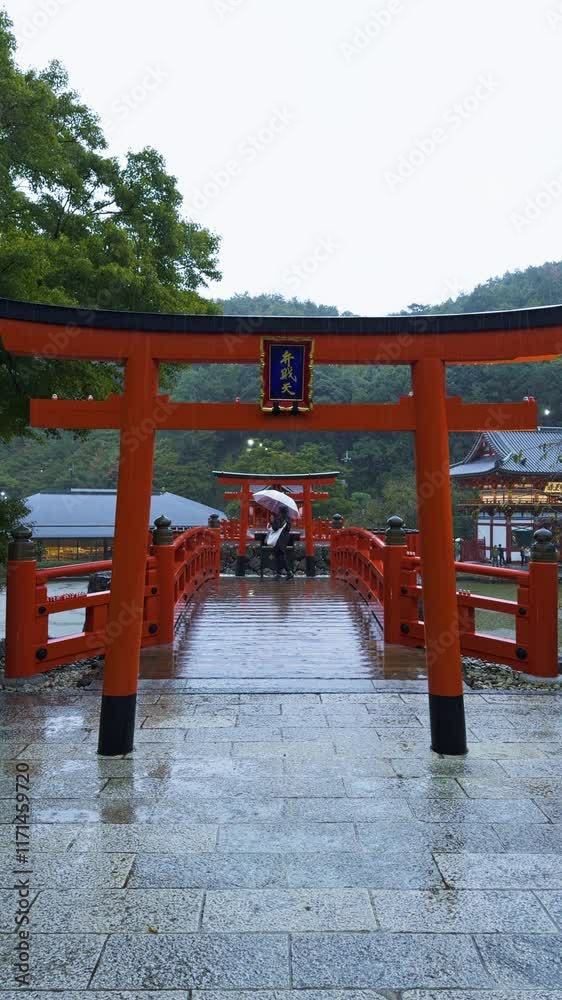 A scenic view of a red Torii gate, wooden bridge, and temple in rainy Osaka-Katsuoji