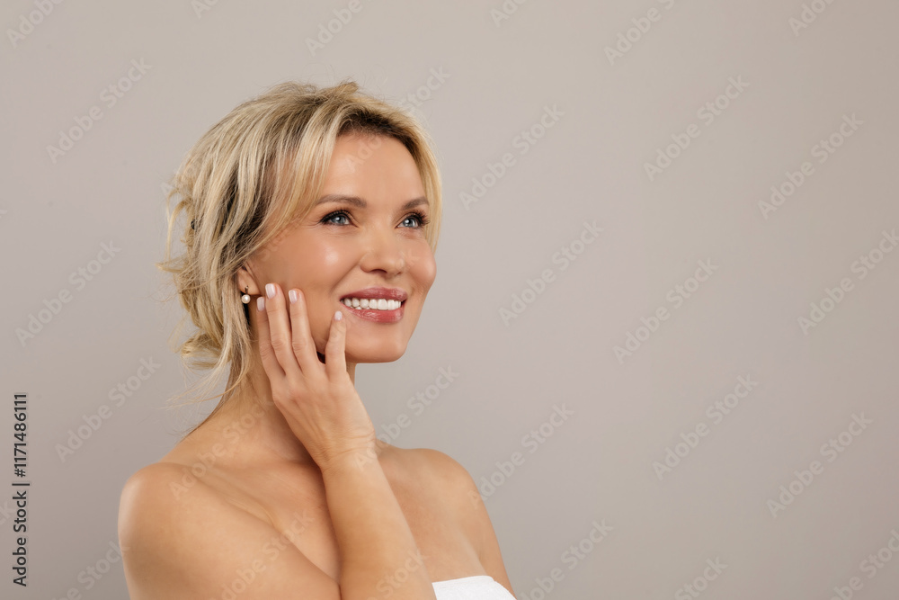 Beautiful middle aged lady wrapped in towel touching her cheek and looking away, smiling attractive mature woman enjoying skincare treatments, standing over grey studio background, copy space. High Beautiful middle aged lady wrapped in towel touching her cheek and looking away, smiling attractive mature woman enjoying skincare treatments, standing over grey studio background, copy space. High