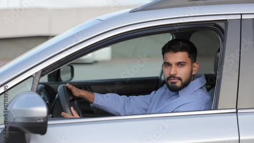 Serious bearded man driving car along street, looking through open window. Side view of handsome brunette male driver in blue shirt looking at camera, while driving vehicle. Driving, lifestyle concept