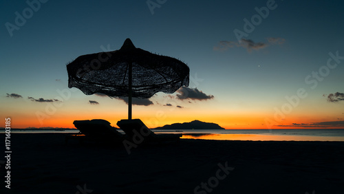 The beginning of the day at the Red Sea. Dark silhouettes of a chaise lounge and an umbrella can be seen on the beach with the orange light from the first rays of the sun and the island of Tiran behin photo