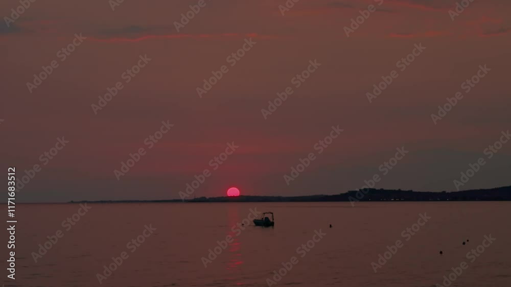 Sunset over Aegean Sea with glowing red sun, calm water and small boat drifting near horizon in Greece. Sunset over Aegean Sea with glowing red sun, calm water and small boat drifting near horizon in Greece.