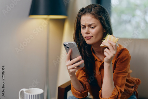 Young woman expressing confusion while looking at her phone, holding a sandwich in a cozy indoor setting, ideal for lifestyle related content focusing on modern communication photo