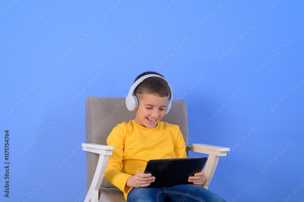 Portrait of happy boy in headphones using digital tablet while sitting on armchair over blue background Portrait of happy boy in headphones using digital tablet while sitting on armchair over blue background