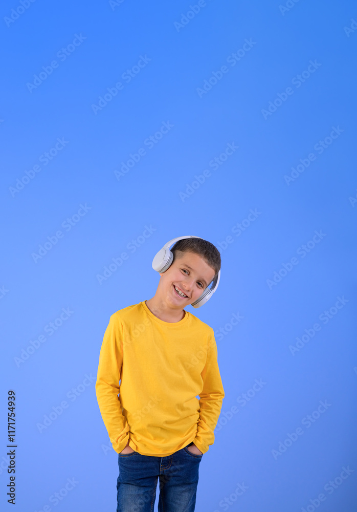 Portrait of cute boy with hands in pockets smiling and listening music over headphones on blue background Portrait of cute boy with hands in pockets smiling and listening music over headphones on blue background