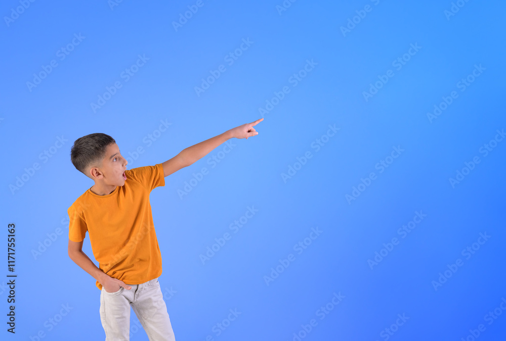 Surprised boy with mouth open pointing finger at copy space while standing against blue background Surprised boy with mouth open pointing finger at copy space while standing against blue background