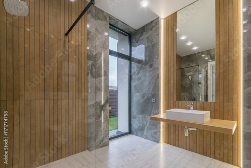 Bathroom with an exclusive design in a luxurious mansion. The room with a shower behind a glass partition is decorated with gray marble tiles and wooden panels.