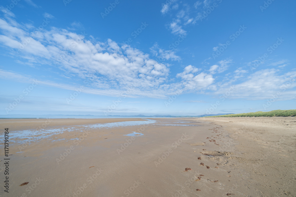 Expansive view of scenic Harlech Beach dunes and grass and patterns in sand to waters edge and horizon under blue sky with high wind-blown white cloud.