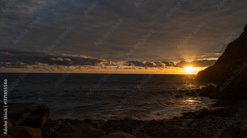 Obraz premium A dramatic sunset over the ocean, with golden light reflecting on the water and dark rocky cliffs framing the scene. A serene and atmospheric coastal landscape.