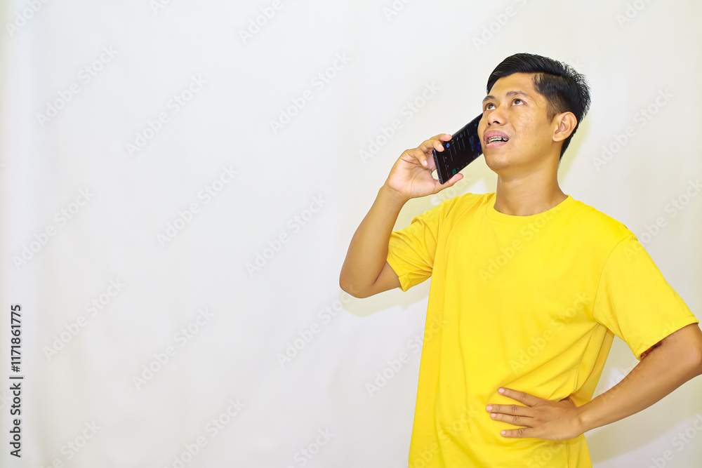A man in a yellow t-shirt, with braces, looking upward while holding a phone to his ear, with his other hand on his waist in a white isolated studio, A man in a yellow t-shirt, with braces, looking upward while holding a phone to his ear, with his other hand on his waist in a white isolated studio,