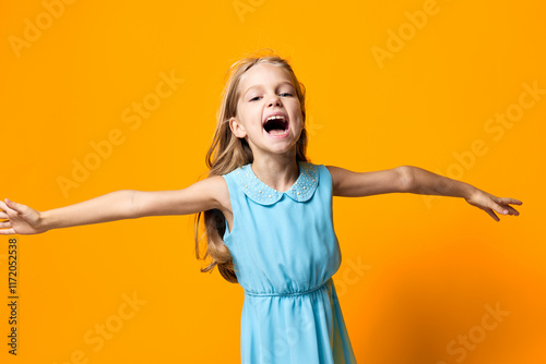 Joyful little girl in a blue dress spreading her arms wide against a vibrant orange background