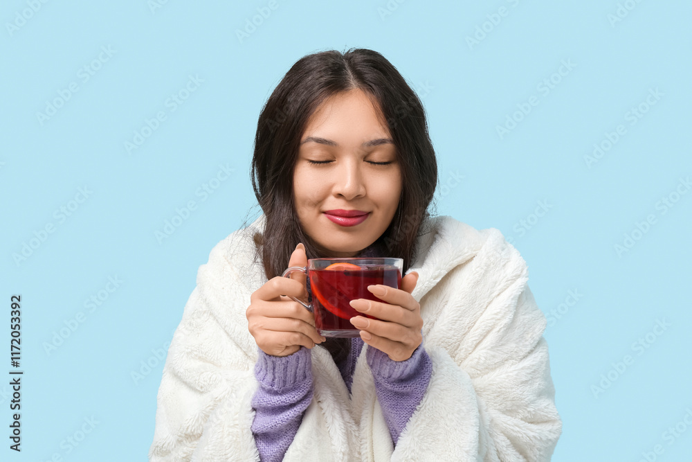 Beautiful young happy Asian woman with cup of hot mulled wine and warm plaid on blue background Beautiful young happy Asian woman with cup of hot mulled wine and warm plaid on blue background