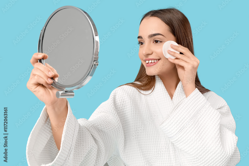Young woman with cotton pad looking in mirror on blue background Young woman with cotton pad looking in mirror on blue background