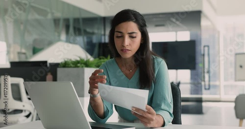 Latina woman employee sit at desk in office, hold papers, read negative news, feels frustrated about project rejection, lay-offs, market decline or partnership termination, fired or position demotion